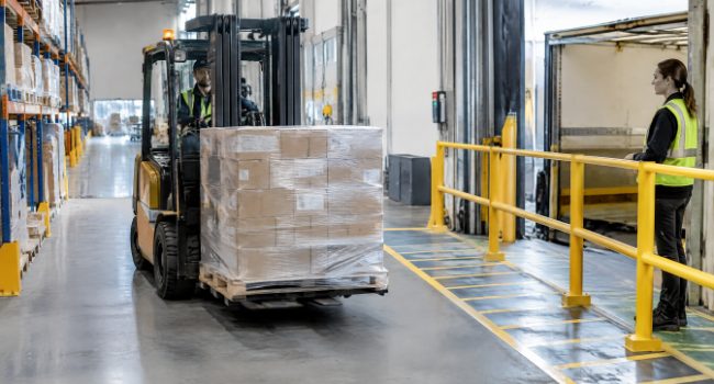 Forklift moving a pallet past a worker standing behind a yellow pedestrian safety barrier