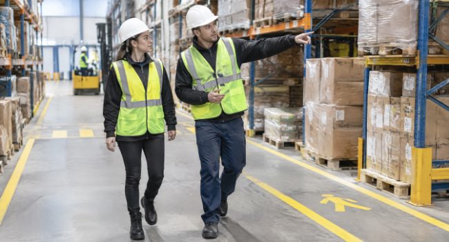 Experienced worker showing a new temporary worker the marked pedestrian walkways in a warehouse aisle